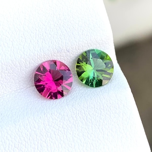Könnte beinhalten: Zwei runde Edelsteine, einer rosa und einer grün, werden auf einer weißen Oberfläche präsentiert. Die Edelsteine sind facettiert und haben eine glänzende Oberfläche.