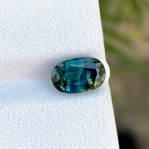 May include: Oval-shaped green gemstone with a blue hue, set on a white background.