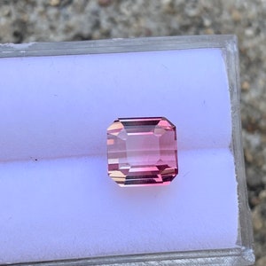 May include: A pink and clear gemstone with a rectangular shape, set in a white display box.
