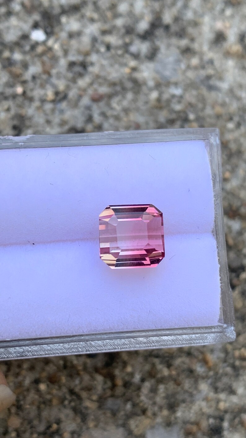 May include: A pink and clear gemstone with a rectangular shape, set in a white display box.
