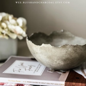May include: A textured, light gray concrete bowl with an irregular, jagged rim. The bowl rests on a stack of three books titled Fix Your Focus, It Is Well, and Worthy. Text at the top reads BLUSH & CHARCOAL and WEE.BLUSHANDCHARCOAL.ETSY.COM.