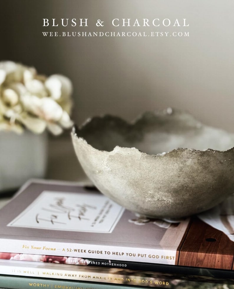 May include: A textured, light gray concrete bowl with an irregular, jagged rim. The bowl rests on a stack of three books titled Fix Your Focus, It Is Well, and Worthy. Text at the top reads BLUSH & CHARCOAL and WEE.BLUSHANDCHARCOAL.ETSY.COM.