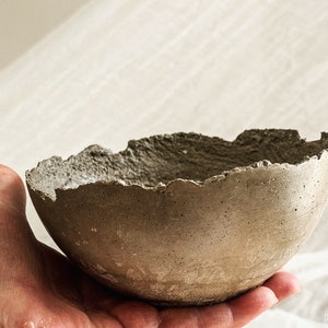 May include: A small, round, gray concrete bowl with a rough, uneven, and jagged rim. The exterior surface of the bowl has a porous, textured appearance with visible small air bubbles and a matte finish, showcasing a raw and industrial aesthetic.
