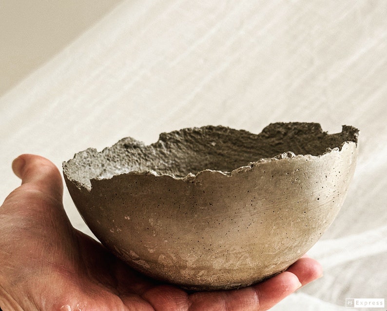 May include: A small, round, gray concrete bowl with a rough, uneven, and jagged rim. The exterior surface of the bowl has a porous, textured appearance with visible small air bubbles and a matte finish, showcasing a raw and industrial aesthetic.