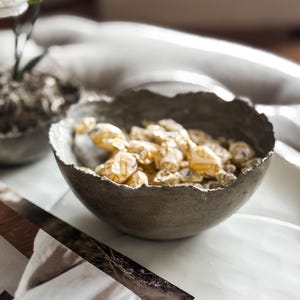 May include: A dark gray, round bowl with a rough, uneven, textured rim. The bowl is filled with multiple small, individually wrapped candies featuring yellow and white packaging. The bowl has a matte finish and a deep, curved basin shape.