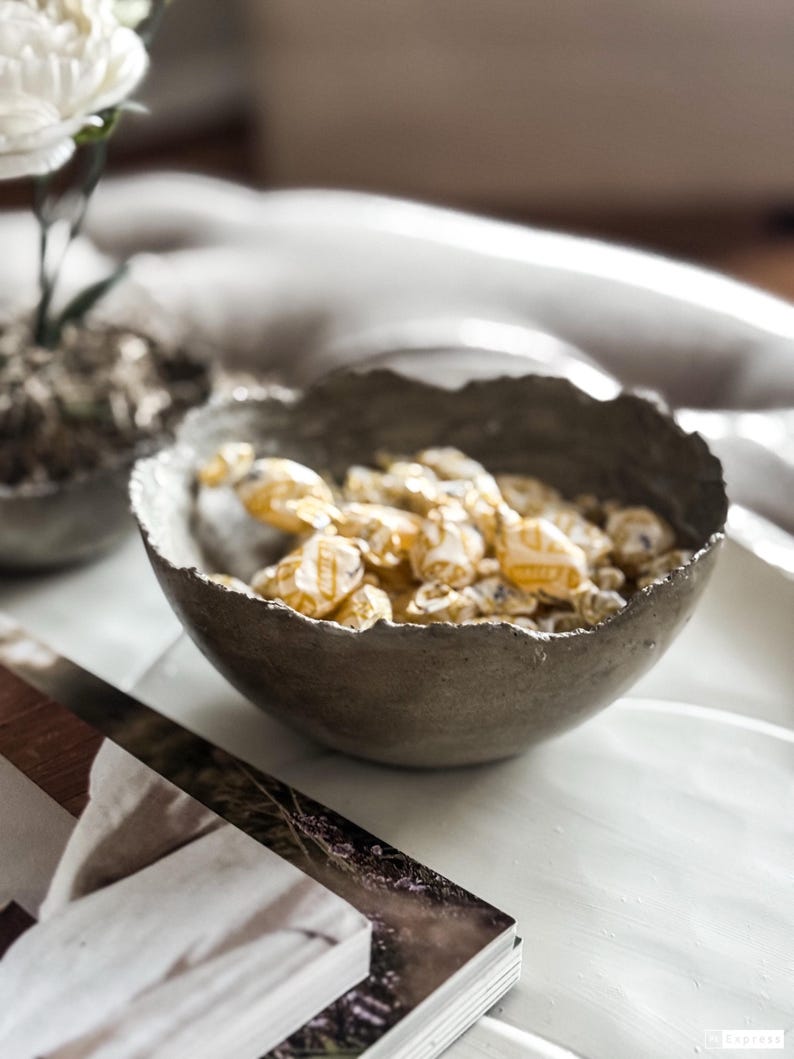 May include: A dark gray, round bowl with a rough, uneven, textured rim. The bowl is filled with multiple small, individually wrapped candies featuring yellow and white packaging. The bowl has a matte finish and a deep, curved basin shape.
