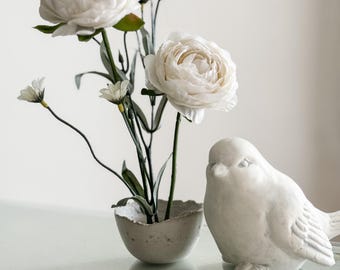 May include: White ceramic bird figurine with a delicate design, sitting on a glass table next to a concrete vase with white flowers.