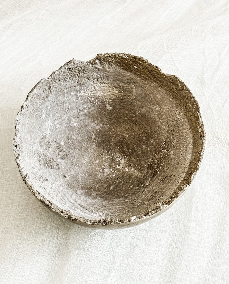 May include: A circular bowl with a rough, uneven, and textured rim. The interior surface features a mottled, earthy brown and grey finish with visible granular details, creating a weathered appearance across the entire concave structure.