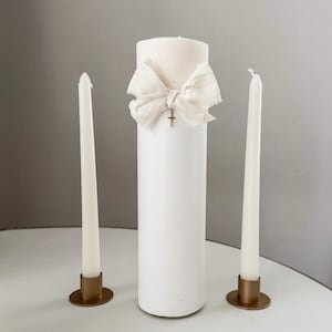 May include: A white pillar candle with a cream-colored fabric bow and a small gold cross pendant. Two white taper candles are on either side, set in gold-colored metal holders. The candles are arranged on a white surface.