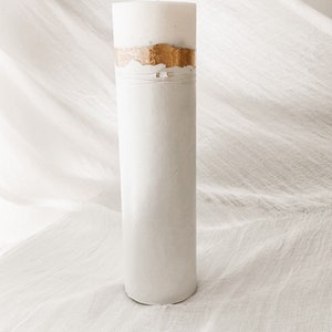 May include: A white pillar candle with a gold band around the middle. The candle is sitting on a white fabric surface.