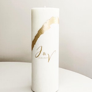 May include: White pillar candle with a gold leaf design and the initials "J & V" and the date "5.28.2023" in gold lettering.