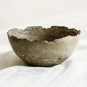 May include: A rough, textured, gray concrete bowl with a slightly uneven rim. The bowl is sitting on a white fabric surface.