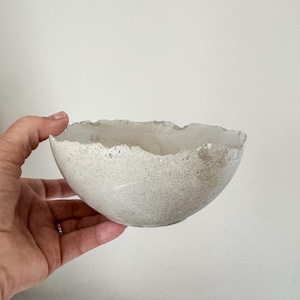 May include: A small, round bowl made of light gray concrete with a rough, uneven, and jagged rim. The exterior surface of the bowl has a porous, speckled texture, while the interior appears smoother with a consistent light gray color throughout.