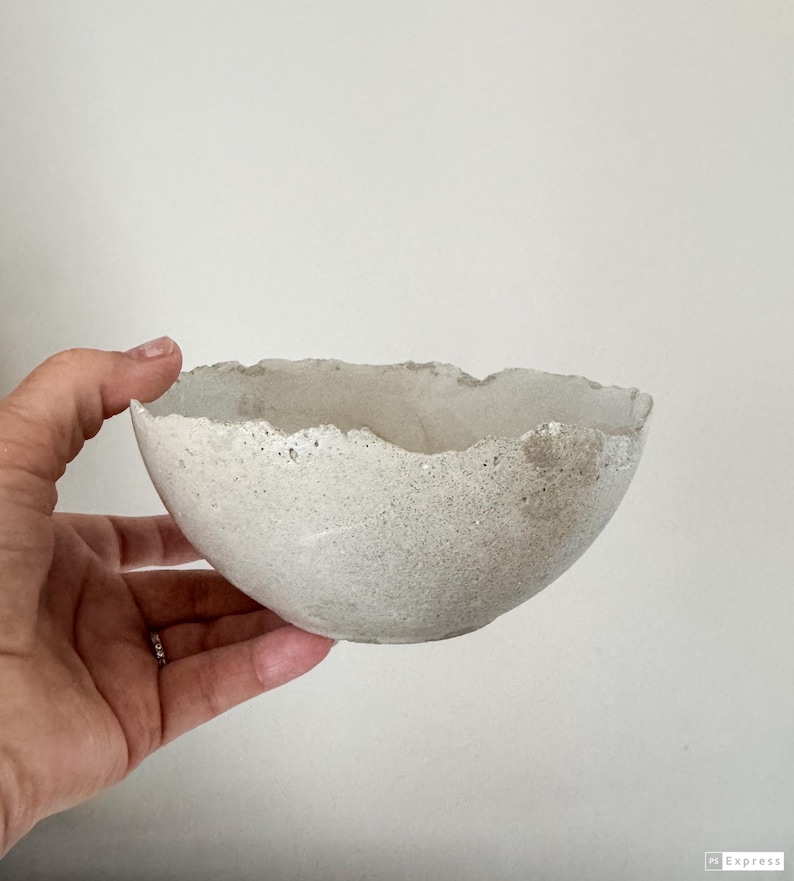May include: A small, round bowl made of light gray concrete with a rough, uneven, and jagged rim. The exterior surface of the bowl has a porous, speckled texture, while the interior appears smoother with a consistent light gray color throughout.