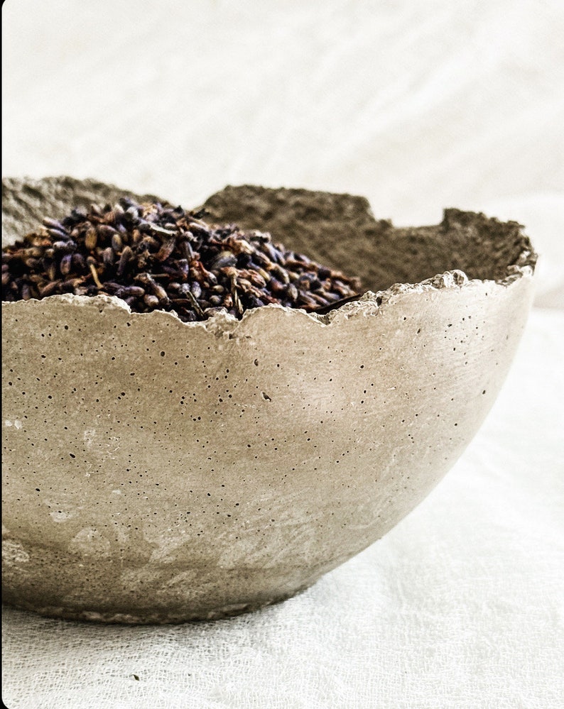 May include: A bowl made of gray concrete with a porous, textured surface and a jagged, uneven rim. The bowl is filled with a mound of dried purple lavender buds, which are clustered together in the center of the container.