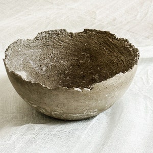 May include: A round, bowl-shaped object with a rough, textured surface and a jagged, uneven rim. The exterior and interior of the bowl feature a mottled, earthy gray-brown color with visible small pores and a coarse, granular finish throughout.