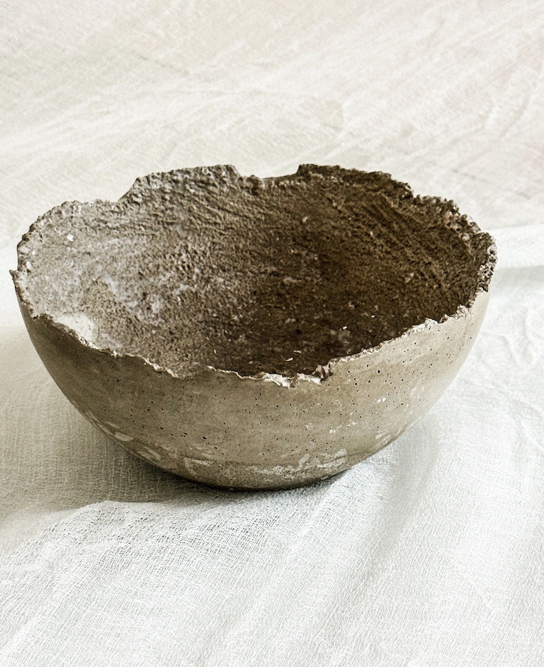 May include: A round, bowl-shaped object with a rough, textured surface and a jagged, uneven rim. The exterior and interior of the bowl feature a mottled, earthy gray-brown color with visible small pores and a coarse, granular finish throughout.