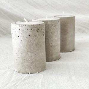 May include: Three gray concrete candle holders with white candles. The candle holders have a rough, textured surface.