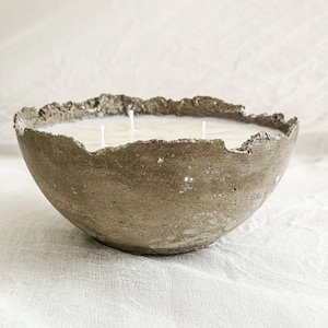 May include: A three-wick candle housed in a bowl-shaped concrete vessel with a rough, uneven, and textured rim. The candle wax is off-white and fills the interior of the gray, porous concrete container, which features a natural, weathered appearance.