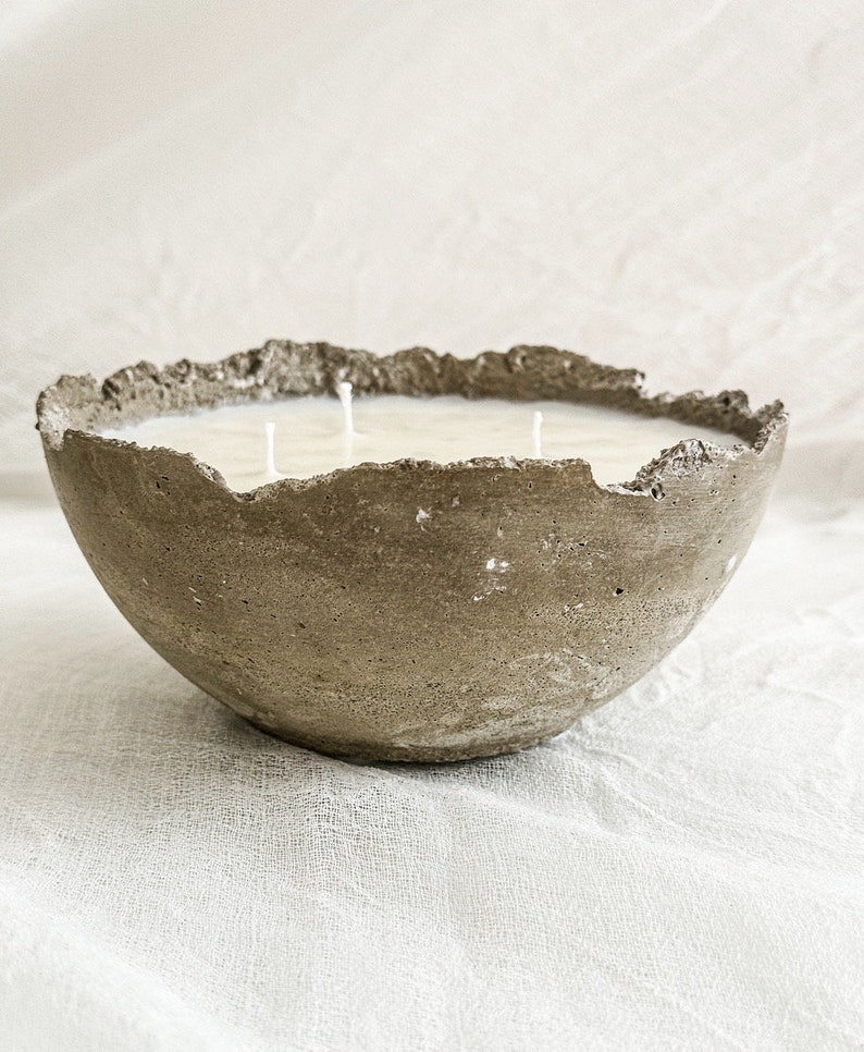 May include: A three-wick candle housed in a bowl-shaped concrete vessel with a rough, uneven, and textured rim. The candle wax is off-white and fills the interior of the gray, porous concrete container, which features a natural, weathered appearance.