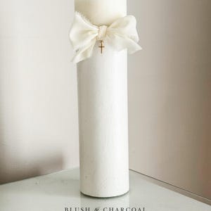 May include: A tall, white pillar candle adorned with a cream-colored bow and a small gold cross pendant. The candle has a textured surface and is presented on a reflective surface. The bottom of the image displays the text "BLUSH & CHARCOAL" and a website address.