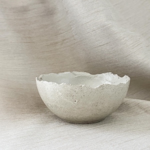 May include: A small, round, off-white bowl with a textured, speckled surface and an uneven, jagged rim. The bowl has a matte finish and a porous appearance, showcasing a handcrafted aesthetic with visible irregularities along the top edge.