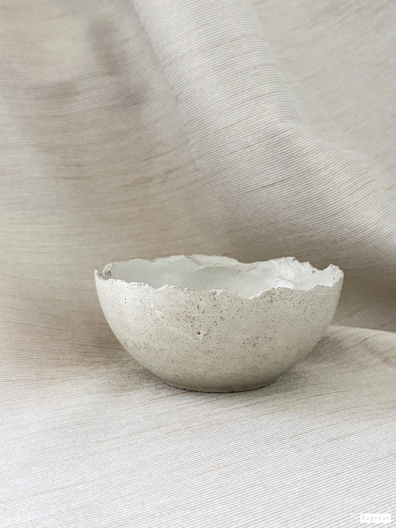 May include: A small, round, off-white bowl with a textured, speckled surface and an uneven, jagged rim. The bowl has a matte finish and a porous appearance, showcasing a handcrafted aesthetic with visible irregularities along the top edge.