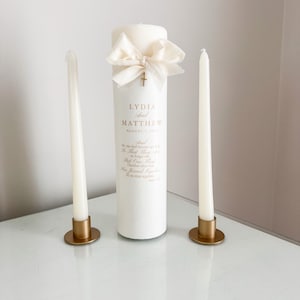 May include: A set of three ivory candles. A large pillar candle is adorned with a cream-colored bow and a small gold cross. Two slender taper candles sit in gold-colored holders. The pillar candle has text that reads "LYDIA and MATTHEW August 1, 2024".