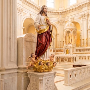 May include: A statue of Jesus Christ in a church setting. The statue features a white robe, a red drape, and a golden sash. Small cherubs surround the base of the statue, which is set on a decorative pedestal.
