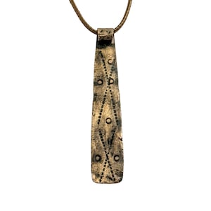 May include: A long, tapered bronze-toned metal pendant suspended from a thin brown cord. The surface features a weathered, textured finish with engraved geometric patterns consisting of intersecting dotted lines and small circular markings.