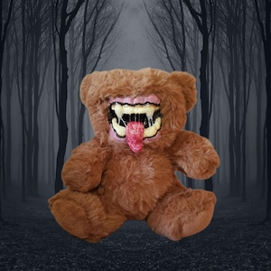 May include: Brown teddy bear with a monstrous mouth featuring sharp teeth and a pink tongue. The bear has a plush texture and is set against a blurred forest backdrop, creating a spooky aesthetic.