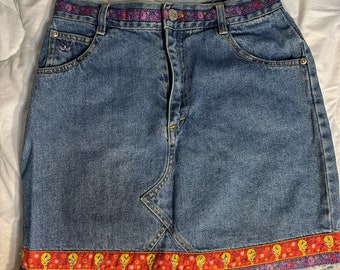 May include: A blue denim skirt with a colorful patterned trim around the waist and hem. The trim features a cartoon character.