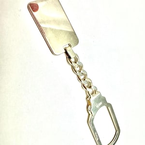 May include: A silver-colored keychain with a rectangular tag and a chain link. The tag has a small red spot on the top left corner. The keychain has a rounded, key-shaped clasp.