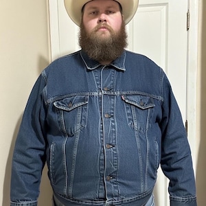 May include: A denim jacket with two chest pockets and button closures. The jacket is paired with light-wash jeans and a cowboy hat with a black crown and a tan brim. The hat has decorative stitching.