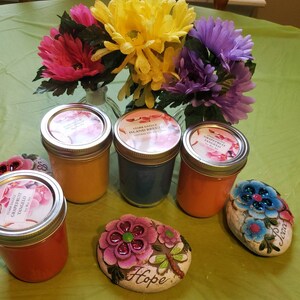 May include: Four glass jars with lids containing colorful candles. The candles are labeled with the scents "Grapefruit Tango", "Island Breeze", and "Star Dazzle". The jars are sitting on a green table with two decorative stones that say "Hope" and "Peace".