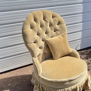 May include: A cream velvet armchair with a button-tufted back and a frilled skirt. The chair has a matching cushion and is sitting on a concrete surface.
