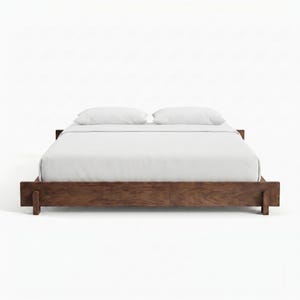 May include: A contemporary wooden bed frame with a low profile. The bed has a dark brown finish, a white fitted sheet, two white pillows, and a white duvet. The bed frame is made of solid wood and has a minimalist design.