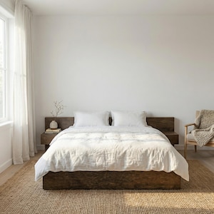 May include: A bedroom with a low, dark wood bed frame, dressed with a white linen duvet and pillows. A woven rug and a wooden bedside table with a vase and book enhance the room's neutral, calming style. A chair with a blanket is in the background.