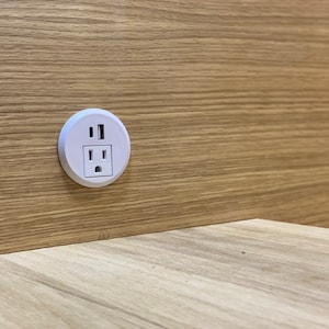 May include: A white electrical outlet with two USB ports is mounted on a light brown wooden surface. The outlet is circular and recessed into the wood. The wood grain is visible.