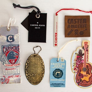 May include: Assortment of clothing tags with various designs.  One tag features a blue and red eagle with the text "CAROCHE LUXURY PRIDE Remember Lemon Li HIS THE". Another tag features a gold oval with the text "CAROCHE" and a geometric pattern. A third tag features a blue and white design with the text "CAROCHE JEANS FINE ROUEK WEAR GUARANTEED HARD WEAR". A fourth tag features a brown leather tag with the text "CASTER A MUERIE". A fifth tag features a black tag with the text "CASTER JEANS SINCE 1971". A sixth tag features a red and white tag with the text "CASTER ejeans.com". A seventh tag features a colorful guitar with the text "Les jeans gita".