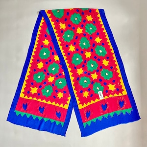 May include: A red, yellow, green, and blue silk scarf with a floral pattern. The scarf has a fringed edge and features a small, white label with the text "YSL".