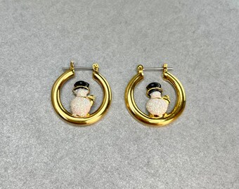 May include: A pair of gold hoop earrings with a snowman charm. The snowman is white with black eyes and a black hat. The snowman is covered in glitter.