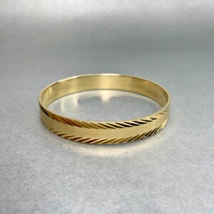 May include: A gold-colored bangle bracelet. The bracelet has a smooth, polished finish with a textured, diagonal pattern around the center. The jewelry is circular and sits on a gray surface.