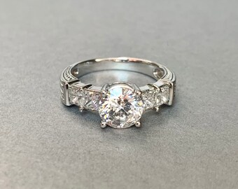 May include: A silver engagement ring with a large round center stone and smaller square stones on the band.