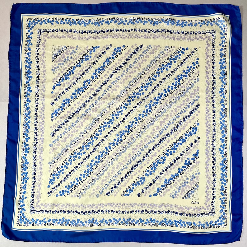 May include: A blue and white patterned silk scarf with a white border and a blue border. The scarf has a diagonal pattern of blue and white flowers. The scarf has the word "Echo" in the bottom right corner.