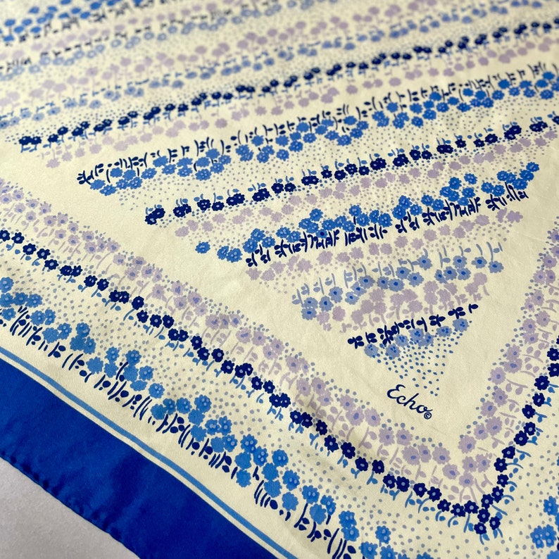 May include: A white silk scarf with a blue and purple floral pattern. The scarf has a blue border and the word "Echo" printed in the center.