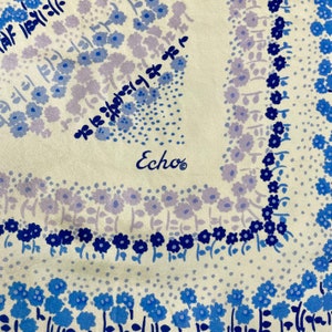 May include: A white silk scarf with a blue and purple floral pattern. The scarf has the word "Echo" printed in the center.