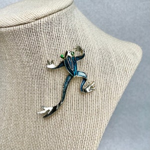 Puede incluir: Un broche de rana de esmalte azul y verde con ojos de pedrería verde. La rana está en una pose de salto con las piernas extendidas.