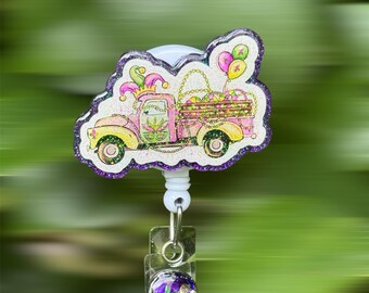 May include: A Mardi Gras themed badge reel featuring a pink and yellow truck carrying balloons and beads. The truck has a glittery finish and is outlined in purple glitter. The badge reel has a clear plastic clip with a purple and green glitter design.