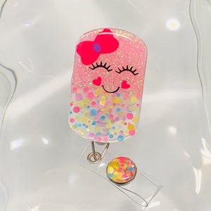 May include: A pink and white glitter badge holder with a smiling face, pink bow, and confetti design. The holder has a clear plastic retractable badge reel with a colourful confetti design.